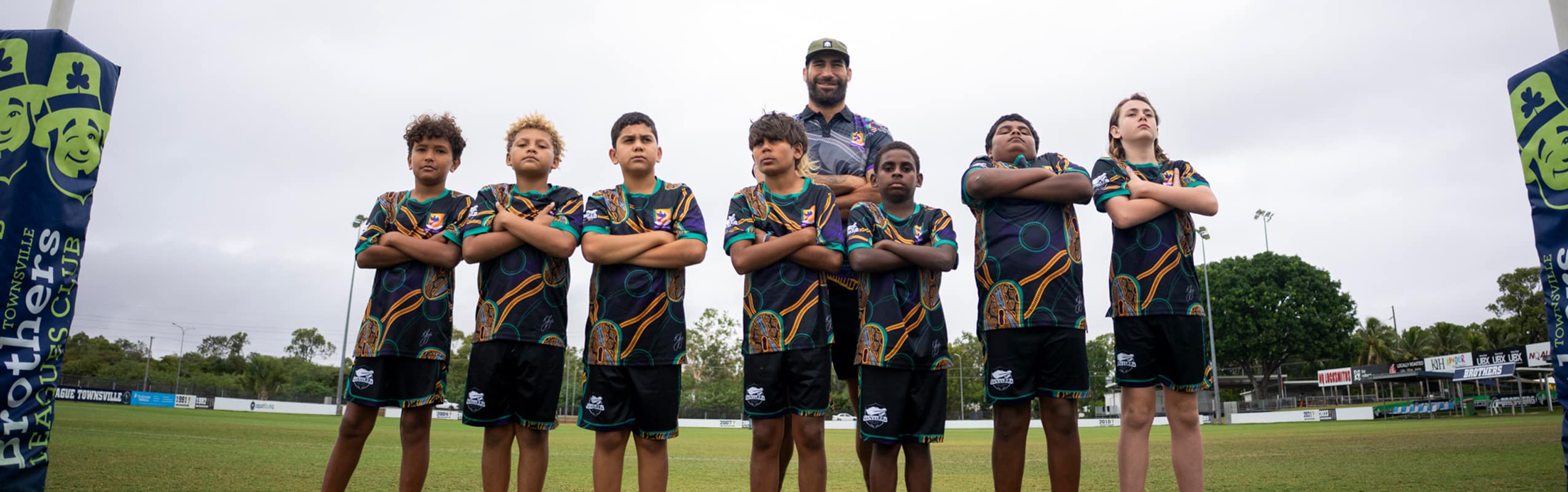 Seven young rugby players in teal and black patterned jerseys stand in a line with arms crossed on a grass field; a coach stands behind them, with banners and bleachers in the background.