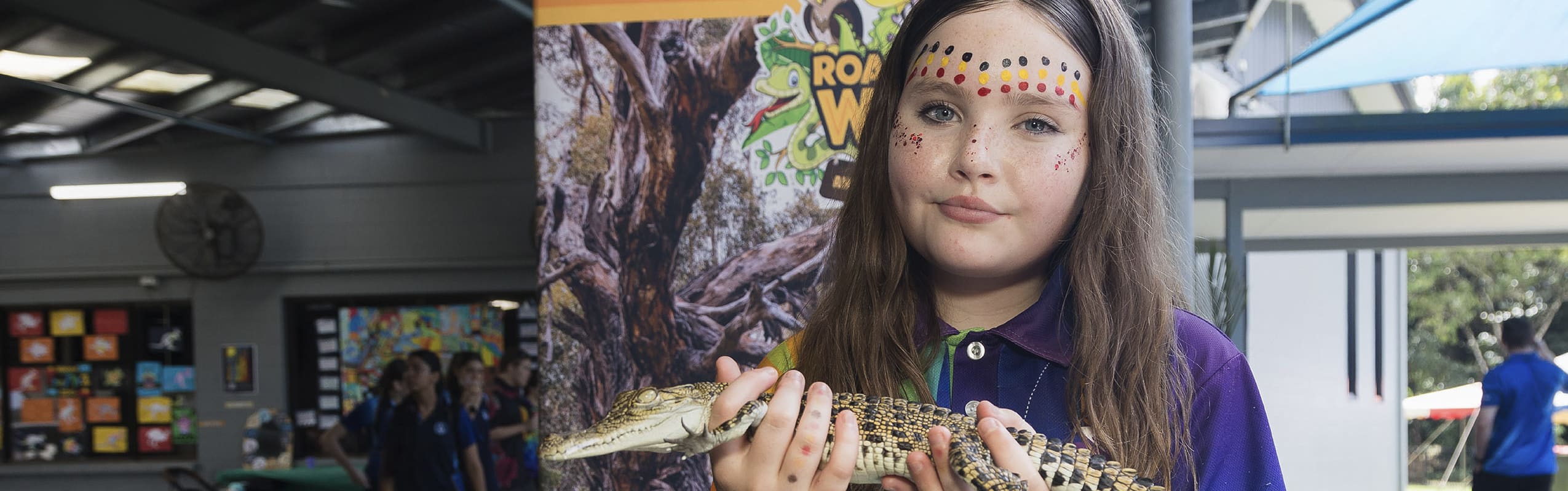 Girl with dotted face paint in a purple shirt holding a small crocodile indoors, smiling at the camera.