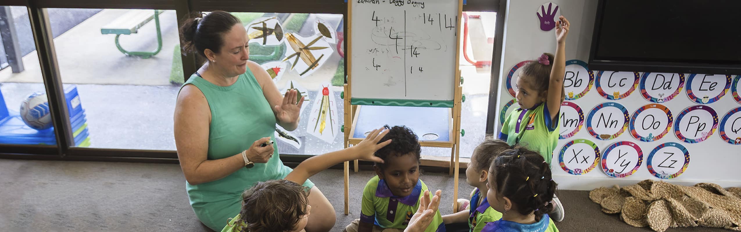 Teacher kneels on the floor with preschoolers during a lesson; a student raises her hand near a whiteboard with an alphabet wall in the background.
